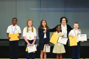Six students stand on stage holding certificates and medals, with an adult standing behind them, posing for a group photo at a math competition.