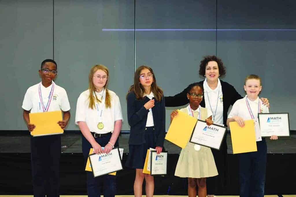 Six students stand on stage holding certificates and medals, with an adult standing behind them, posing for a group photo at a math competition.
