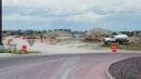 A construction site with dirt piles, vehicles, and orange traffic barrels beside a curved road under a cloudy sky.