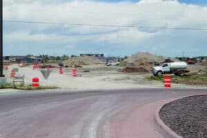 A construction site with dirt piles, vehicles, and orange traffic barrels beside a curved road under a cloudy sky.