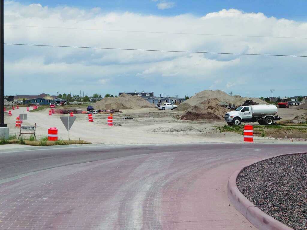 A construction site with dirt piles, vehicles, and orange traffic barrels beside a curved road under a cloudy sky.