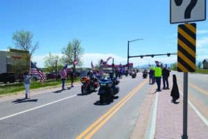 Motorcyclists ride down a street lined with people holding American flags on a sunny day. Traffic and road signs are visible.