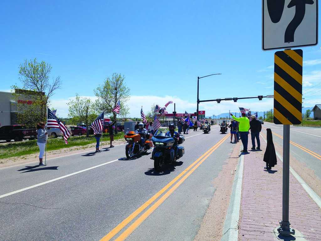 Motorcyclists ride down a street lined with people holding American flags on a sunny day. Traffic and road signs are visible.
