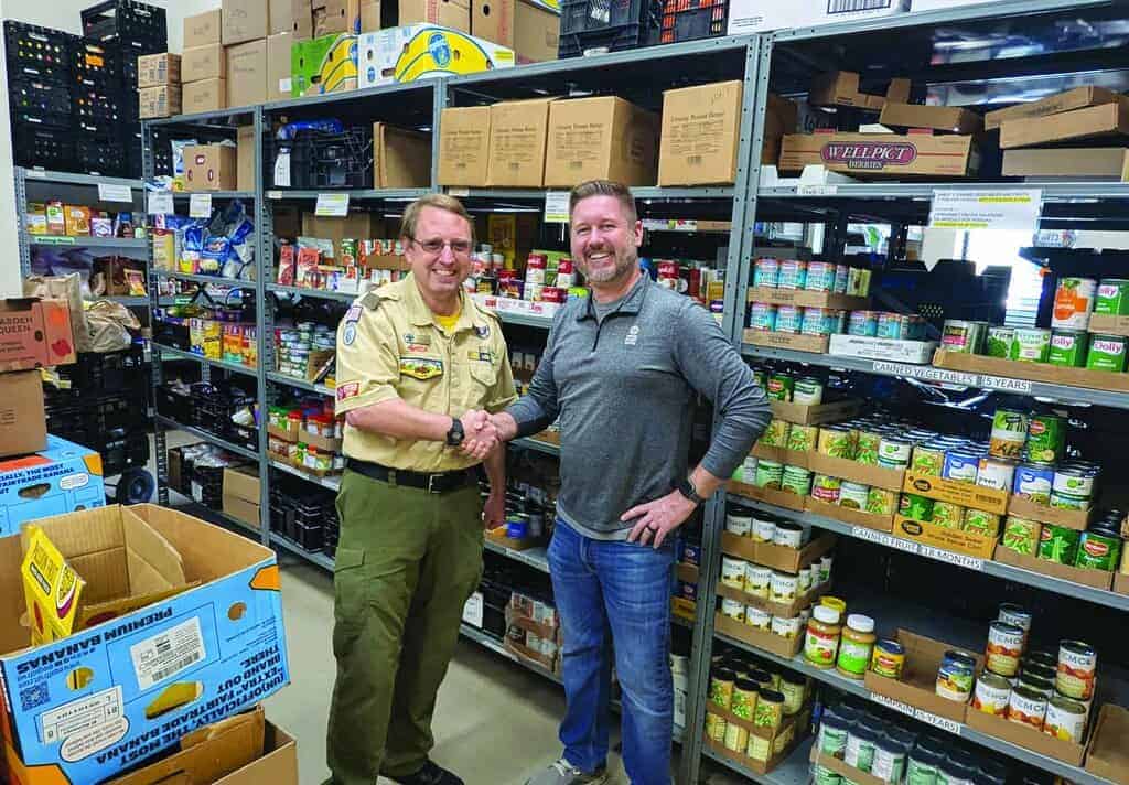 Two men shake hands in front of shelves stocked with canned goods and boxes in a food pantry or storage area.