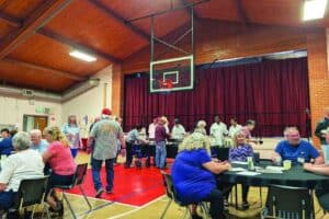 People sit and socialize at tables in a gymnasium with a high wooden ceiling, while others line up near a stage for food service.