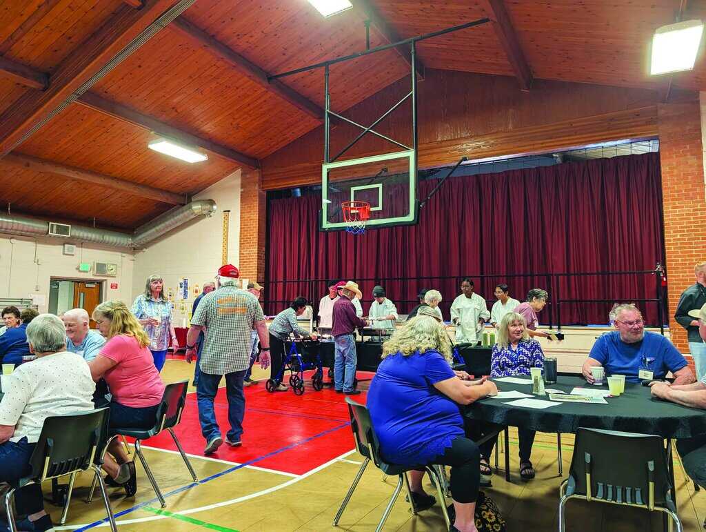 People sit and socialize at tables in a gymnasium with a high wooden ceiling, while others line up near a stage for food service.