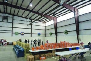 People are gathered in a large indoor hall with hay bales, rows of orange food boxes labeled “Thankful for You,” and tables with various items.