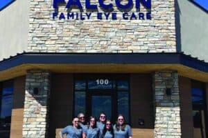 Six people in matching gray shirts stand in front of the Falcon Family Eye Care building, which has a stone facade and prominent blue sign.