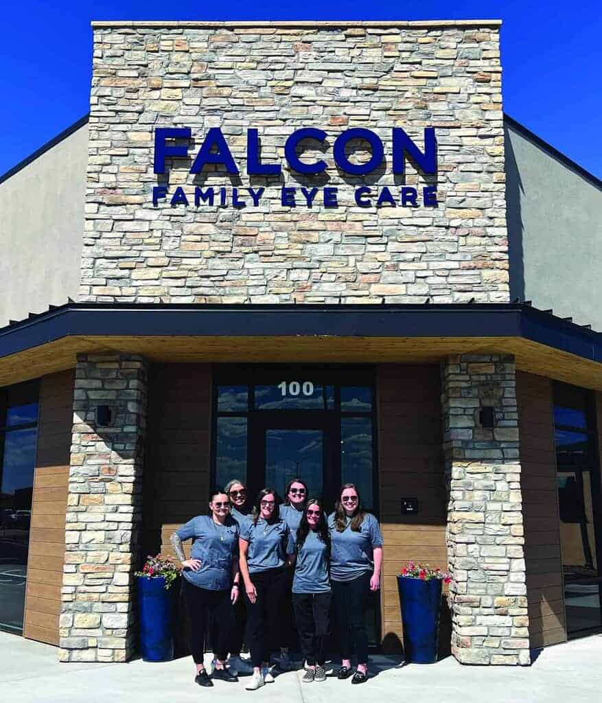 Six people in matching gray shirts stand in front of the Falcon Family Eye Care building, which has a stone facade and prominent blue sign.