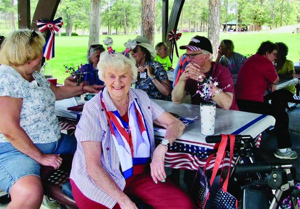 A group of older adults sit at a picnic table decorated with patriotic red, white, and blue items under a pavilion in a park.