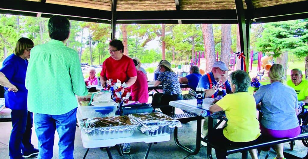 A group of people gather under a pavilion at a park, sitting at picnic tables and serving food from trays on a buffet table.