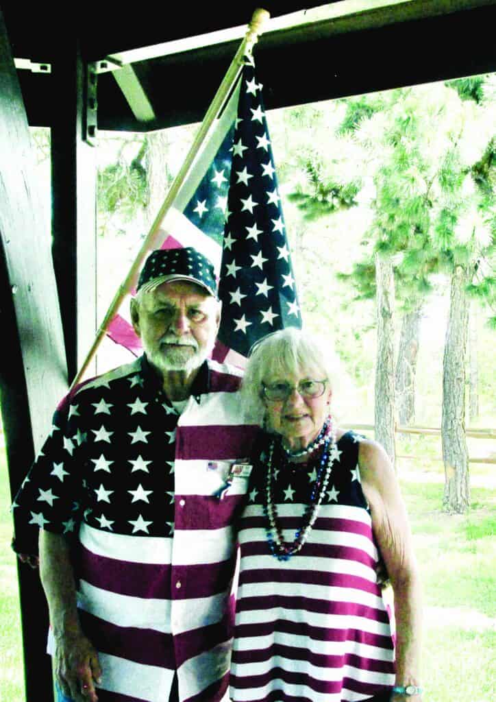 An elderly man and woman stand together in front of a US flag, both wearing American flag-themed clothing, outdoors near trees.