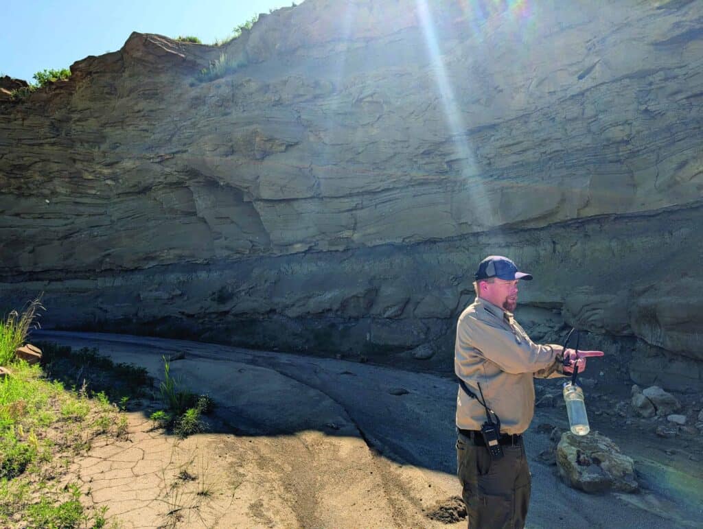 A man in outdoor gear stands and gestures in front of a steep, layered rock formation under direct sunlight.
