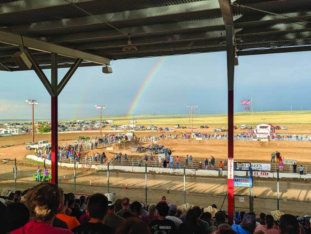Spectators watch a rodeo event at an outdoor arena with a rainbow visible in the cloudy sky and cars parked in the background.