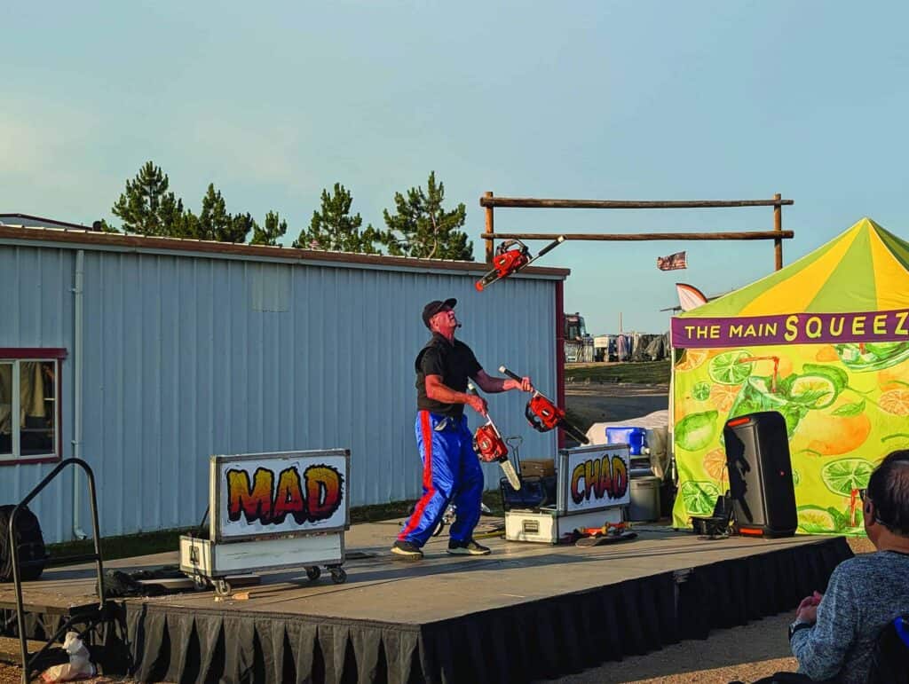 A performer juggles three running chainsaws on an outdoor stage next to a lemon-themed tent and a building, with a small audience watching.