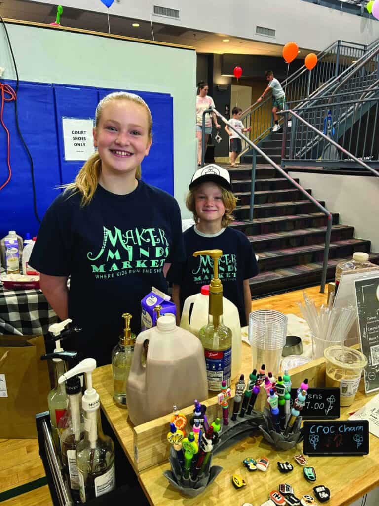 Two children stand behind a drink stand at an indoor market, smiling at the camera. The counter in front of them displays syrups, cups, and a cash box.