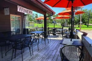 Outdoor patio at a winery with black metal tables and chairs, orange umbrellas, and a wooden deck, surrounded by greenery on a sunny day.