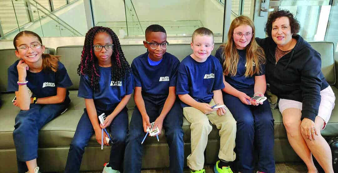 Five children and one adult sit on a couch indoors. The children are wearing matching blue shirts and holding cards or small items.