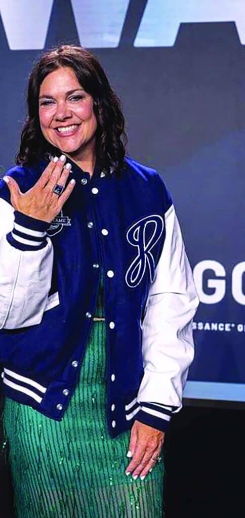 A woman wearing a blue and white varsity jacket and a green dress stands indoors, smiling and showing her hand.