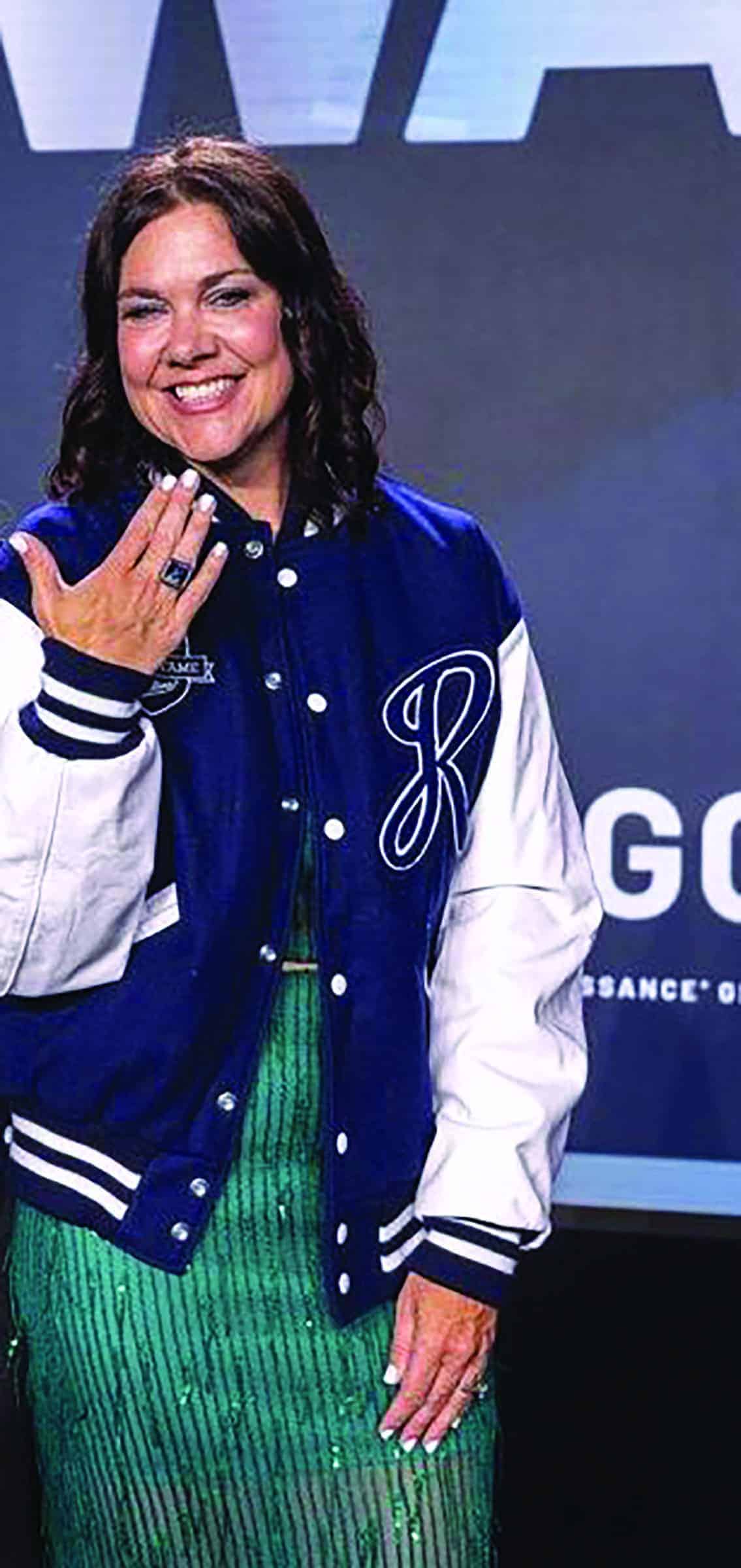 A woman wearing a blue and white varsity jacket and a green dress stands indoors, smiling and showing her hand.