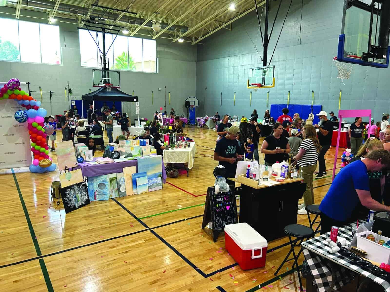 People attend an indoor craft fair in a gymnasium with tables displaying artwork, handmade goods, and decorations. Balloon arrangements and vendors are visible.