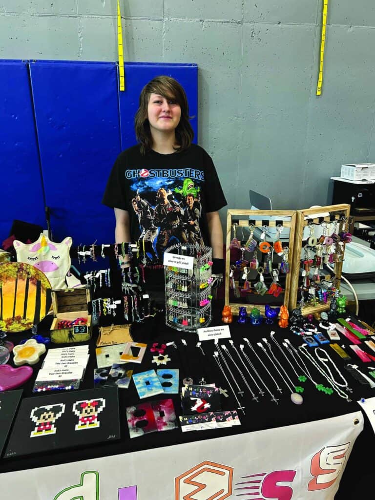 A person stands behind a display table of handmade jewelry, keychains, and crafts at an indoor event, wearing a Ghostbusters T-shirt.