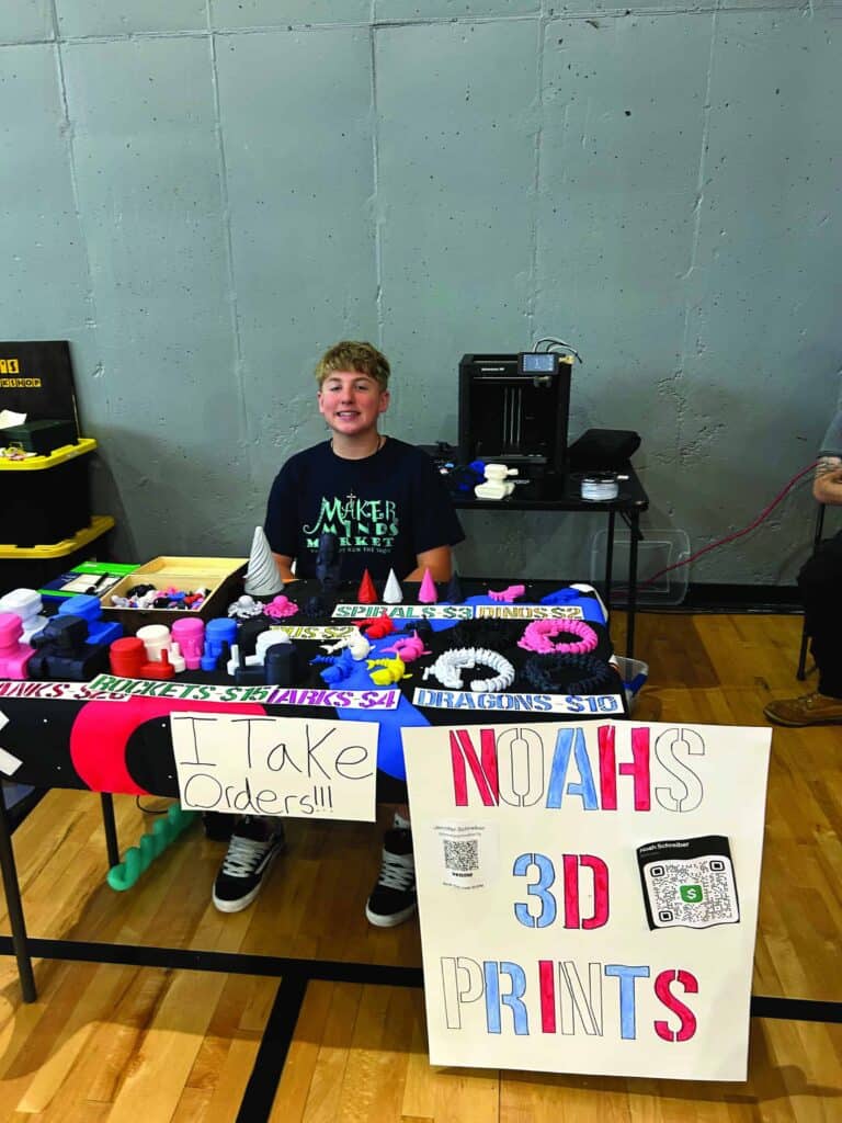 A boy sits behind a table displaying various 3D-printed items and signs that read “I Take Orders!!!” and “Noah’s 3D Prints” at an indoor event.