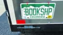 A Colorado license plate on a vehicle reads "BOOKSHP" in green letters, with white mountains in the background.