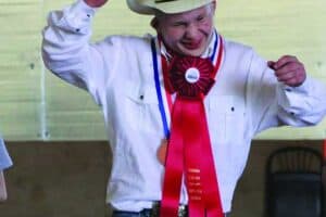 A person wearing a cowboy hat, white shirt, and jeans smiles and raises their arms while displaying medals and a large red ribbon on their chest.