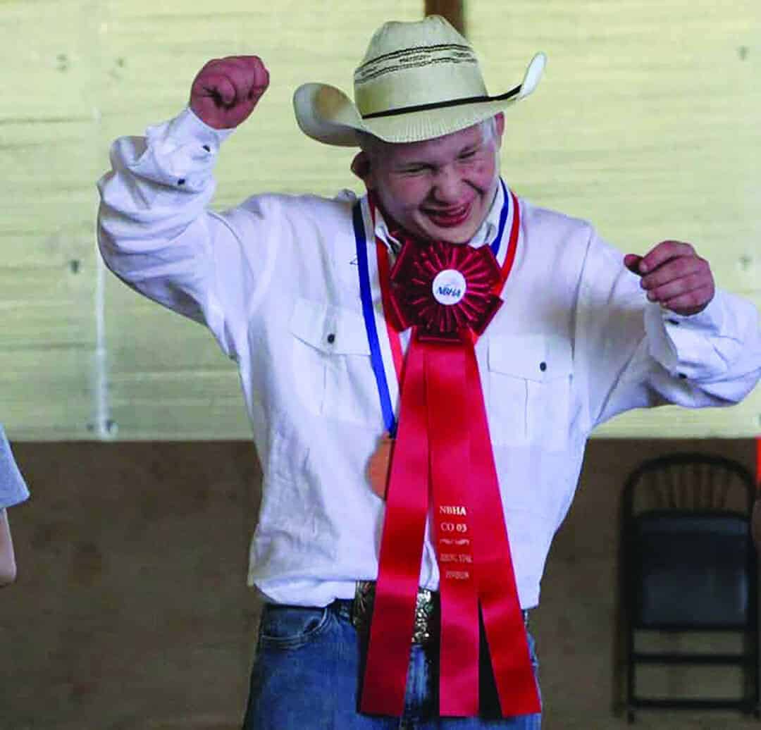 A person wearing a cowboy hat, white shirt, and jeans smiles and raises their arms while displaying medals and a large red ribbon on their chest.