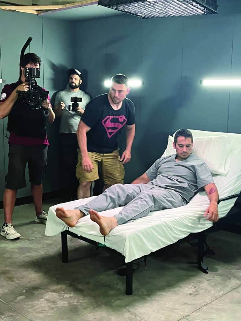 A man in gray clothes sits on a hospital bed while three crew members, one holding a camera, stand nearby in a studio setting.