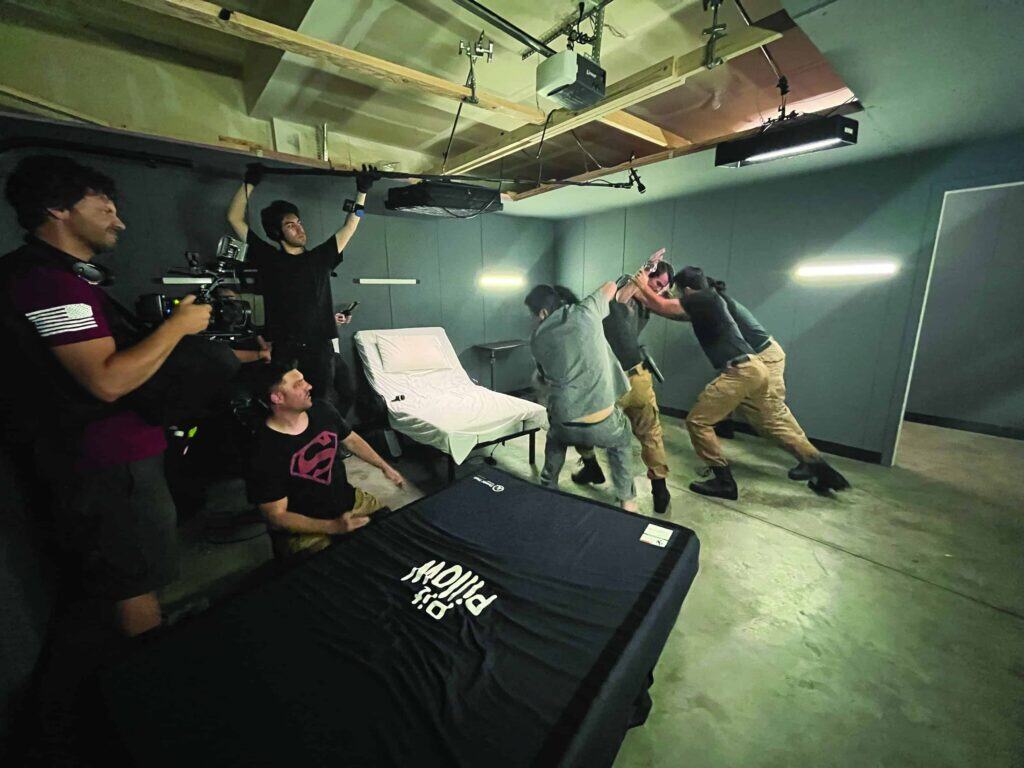 A film crew records two men in tactical pants simulating a fight in a room with gray walls, while others observe and operate camera equipment.