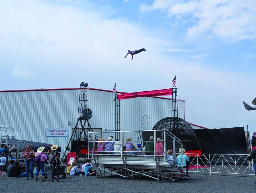 A person is seen flying through the air above a circus setup, while spectators sit on bleachers and watch outdoors near a large building.