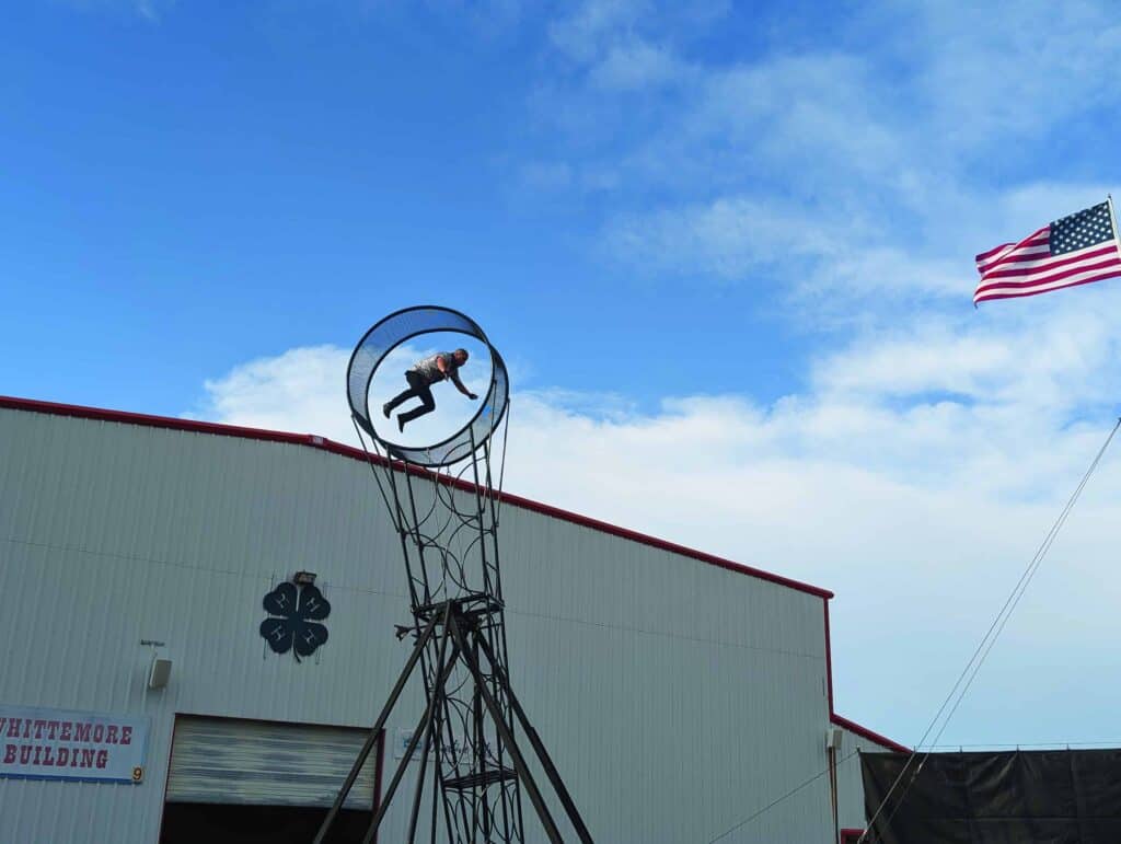 A person is mid-air inside a metal hoop structure in front of a building, with an American flag waving in the sky.