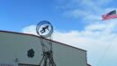 A person is mid-air inside a metal hoop structure in front of a building, with an American flag waving in the sky.