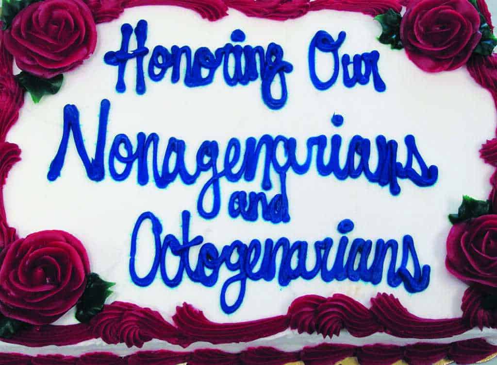 A decorated cake with red roses and blue icing text that reads, "Honoring Our Nonagenarians and Octogenarians.