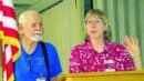 An older man and woman stand at a podium indoors. The man wears a blue shirt and suspenders, while the woman, in a red patterned shirt, gestures with her hand. An American flag is visible.