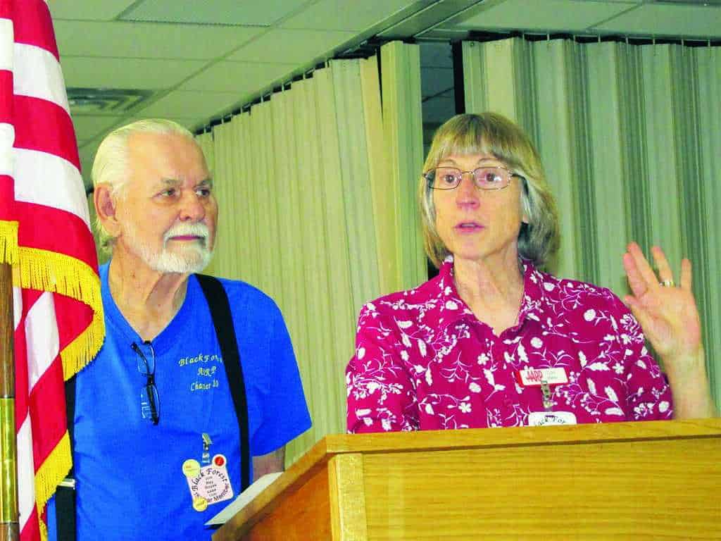 An older man and woman stand at a podium indoors. The man wears a blue shirt and suspenders, while the woman, in a red patterned shirt, gestures with her hand. An American flag is visible.