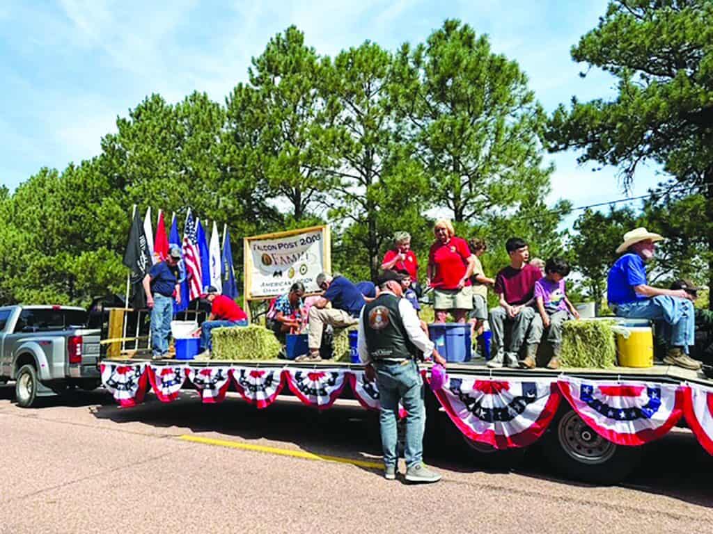 A parade float decorated with red, white, and blue bunting carries people sitting on hay bales, with flags and a banner displayed in the background on a sunny day.