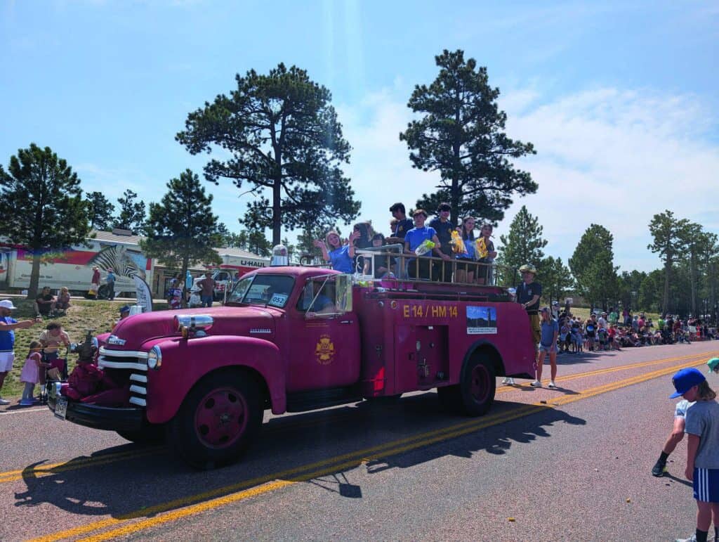 A vintage pink fire truck carrying children drives in a parade, with crowds watching from both sides of the road on a sunny day.