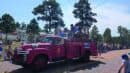 A vintage pink fire truck carrying children drives in a parade, with crowds watching from both sides of the road on a sunny day.