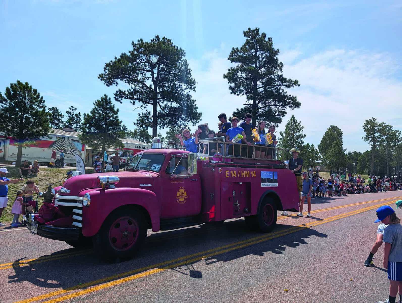 A vintage pink fire truck carrying children drives in a parade, with crowds watching from both sides of the road on a sunny day.