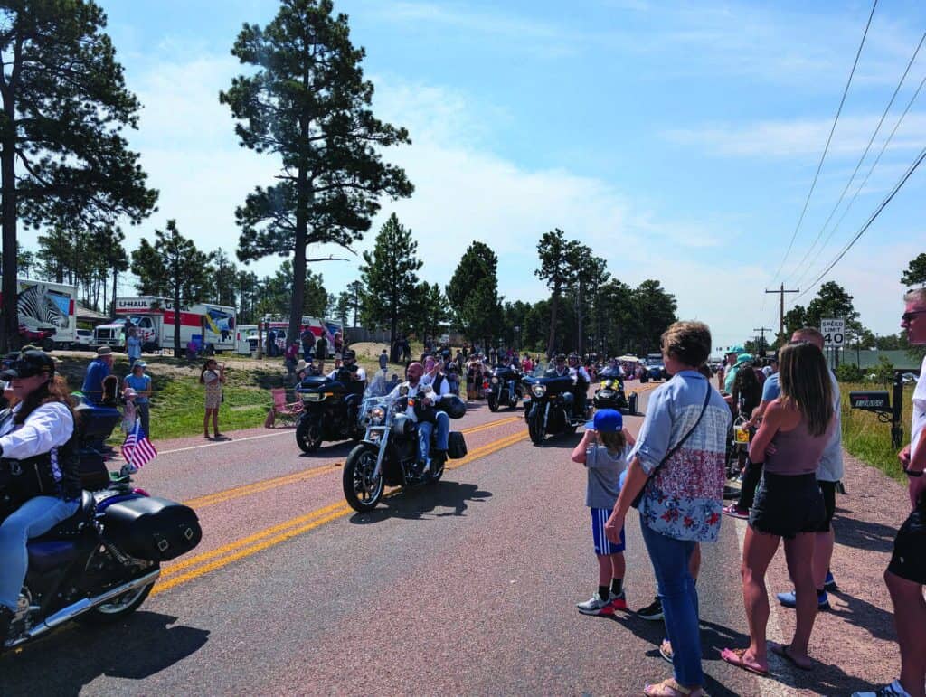 A group of motorcyclists rides down a road lined with spectators, including families and children, on a sunny day with trees and parked vehicles in the background.