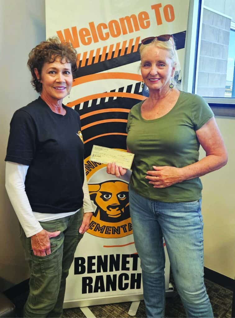 Two women stand indoors holding a check in front of a "Welcome to Bennett Ranch" banner.