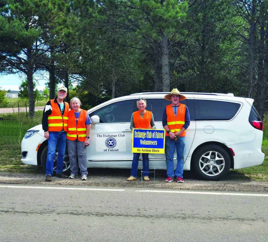 Four adults wearing orange safety vests stand in front of a white van on a roadside, holding a sign that reads "Exchange Club of Falcon Volunteers." Trees and grass are in the background.