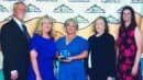 Five people stand together posing for a photo in front of a Lutheran Family Services banner; the woman in the center holds an award.