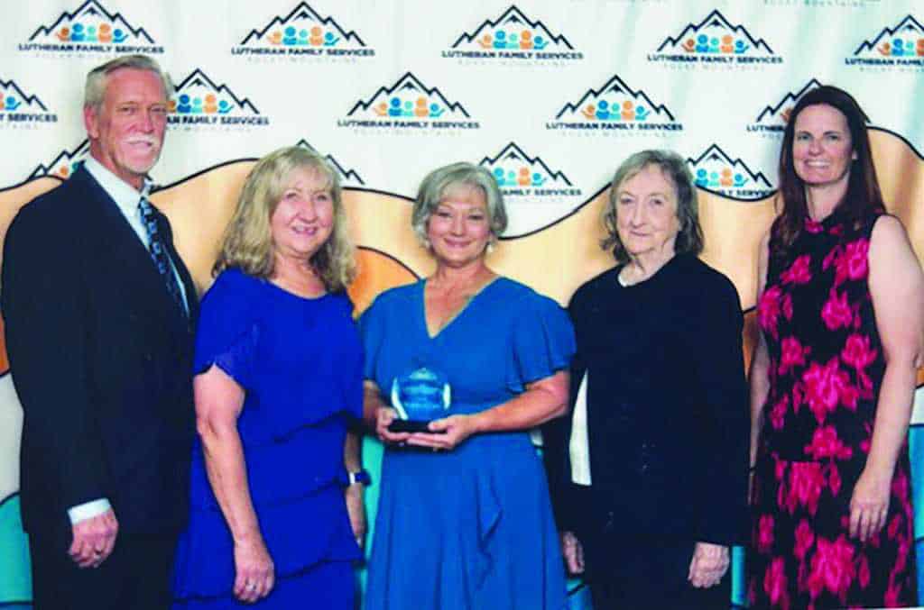 Five people stand together posing for a photo in front of a Lutheran Family Services banner; the woman in the center holds an award.