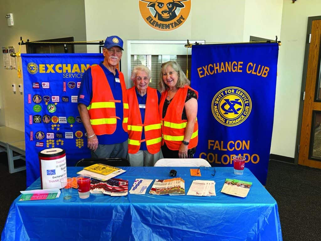 Three adults wearing orange safety vests stand behind an informational table for the Exchange Club of Falcon, Colorado, with banners, pamphlets, and a donation container displayed.