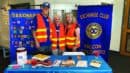 Three adults wearing orange safety vests stand behind an informational table for the Exchange Club of Falcon, Colorado, with banners, pamphlets, and a donation container displayed.