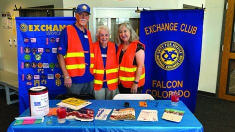 Three adults wearing orange safety vests stand behind an informational table for the Exchange Club of Falcon, Colorado, with banners, pamphlets, and a donation container displayed.